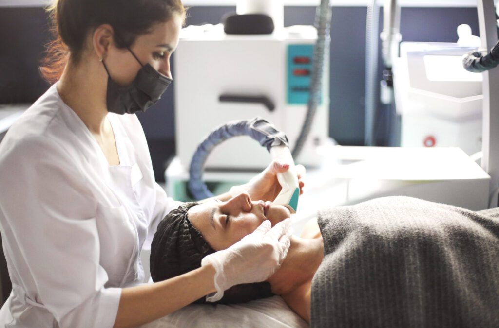 Female Aesthetician in Lahore clinic in mask doing laser therapy procedure on face of relaxed woman 