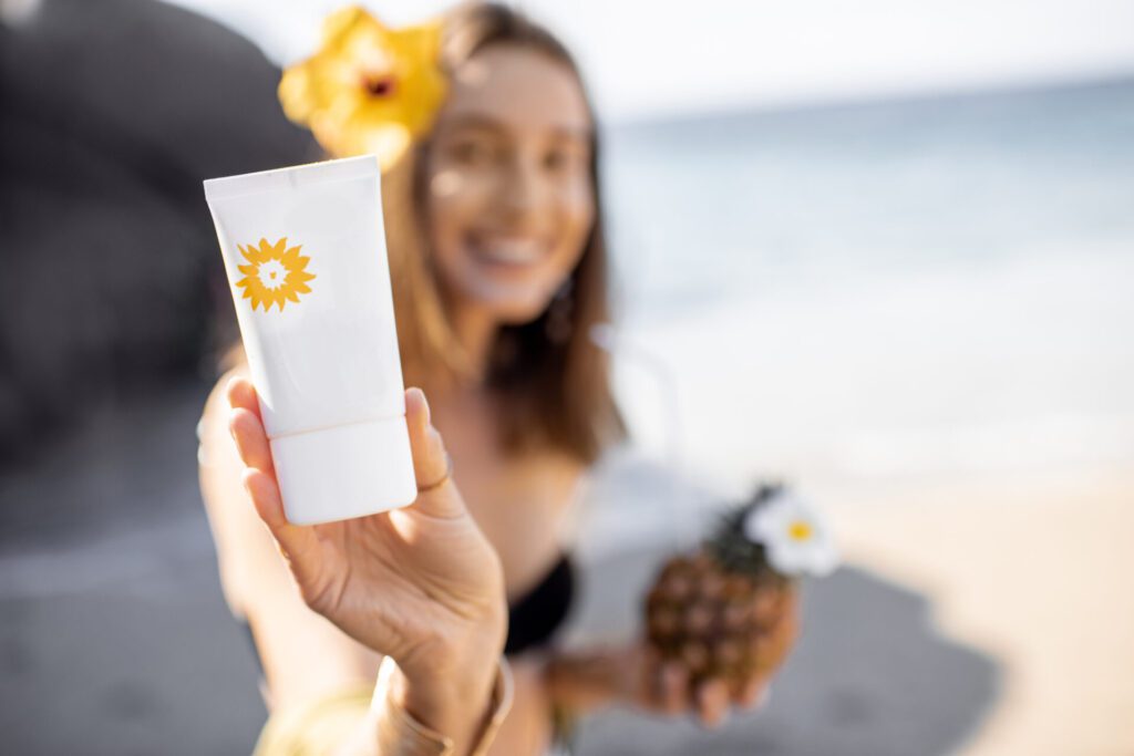 Beautiful smiling girl showing tube with sunscreen lotion while resting on the beach, image focused on the tube. Sun protection concept for skin